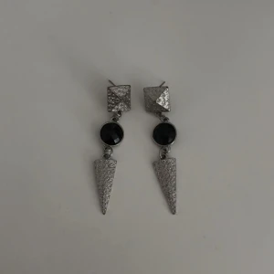 Silvriga örhängen - Snygga örhängen i silverfärg. Örhängena har en fyrkantig topp, rund svart sten i mitten och avslutas med en spetsig, texturerad trekant. Perfekta för att lyfta vilken outfit som helst.