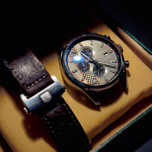 TAG Heuer Carrera Senna kronograf - Säljer en TAG Heuer Carrera Senna kronograf med racinginspirerad urtavla i silver och svart, schackrutigt mönster och röda detaljer. Klockan har en robust boett i stål och ett brunt läderarmband med perforeringar och snyggt spänne med TAG Heuer-logga.