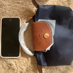Mörkblå axelväska från Longchamp - Snygg mörkblå axelväska från Longchamp med bruna skinnhandtag och detaljer i guld. Väskan har klassisk design, präglad logga på locket och medföljande axelrem. Perfekt storlek för mobilen och det viktigaste. Stängs med dragkedja och knapp.