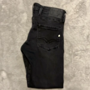 Replay Anbass svarta jeans, stl 30 - Säljer ett par svarta Replay Anbass jeans i slim fit. Jeansen har klassisk femficksdesign, snygga detaljer vid fickorna och en lätt tvättad look. De är tillverkade i stretchig bomullsdenim för extra komfort och har silverfärgade knappar och nitar. Och man ser lite på taggen över fickan med Replay R så är det lite skrynkligt men det blir så efter första tvätten på vissa.