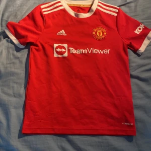 Manchester United matchtröja Adidas 21/22 hemma - Röd Manchester United fotbollströja från Adidas med vita detaljer, klubbmärke på bröstet och TeamViewer sponsortryck. Klassiska tre ränder på axlarna, vit krage och ärmslut. Tillverkad i lätt och ventilerande material, perfekt för träning eller match.
