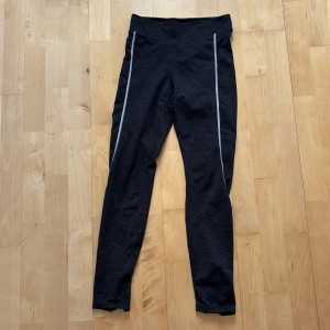 Svarta sportleggings från H&M Sport - Svarta sportiga leggings från H&M Sport i storlek S. De har gråa detaljer längs benen och dragkedjor vid bensluten. Perfekta för träning eller löpning, med en tight passform och stretchigt syntetmaterial som ger bra rörelsefrihet.