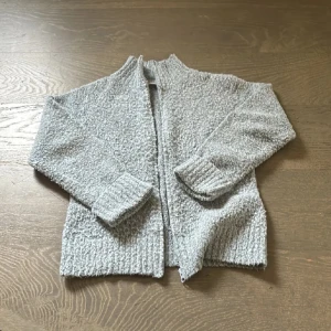 Grå cardigan jacka från Fransa - Mysig grå cardigan jacka från Fransa i storlek S. Jackan är stickad med en fluffig, texturerad yta och har långa ärmar samt ribbade muddar. Perfekt för lager-på-lager och chill dagar. Stängs med dragkedja framtill.