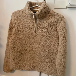 Beige teddy fleece med half zip - Supermjuk och mysig beige teddy fleece. Tröjan har en hög krage och en praktisk half zip-dragkedja framtill. Perfekt för kyliga dagar och har långärmad passform. Oversized och fluffig look som är riktigt trendig just nu.