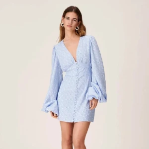 Adoore ljusblå kort klänning med puffärmar strl 36 - Supersöt ljusblå kort klänning med v-ringning, puffiga långärmar och små blommiga detaljer. Klänningen har knappar framtill och markerad midja som ger en snygg siluett. Perfekt för vår och sommar! Strl 36. Helt ny, aldrig använd! Nypris: 1495kr 