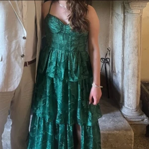 Grön långklänning med volanger - Säljer en grön, lång volangklänning från Azazie. Använd 1 gång på ett bröllop i juni. Litet hål i innertyget vid vänstra bröstet men ingenting som syns och påverkar inte passformen. Går att sy igen lätt om man kan sånt. Köptes för 2200kr