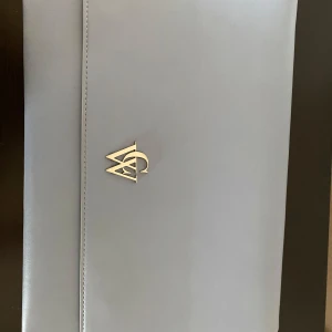 Ljusblå kuvertväska från Carin Wester  - Snygg ljusblå kuvertväska från Carin Wester. Väskan har ett stilrent metallogo framtill och knapplås. Insidan är fodrad med mönstrat tyg med logotryck och har ett praktiskt innerfack. Perfekt för att bära laptop eller viktiga papper. Mått 26x36.