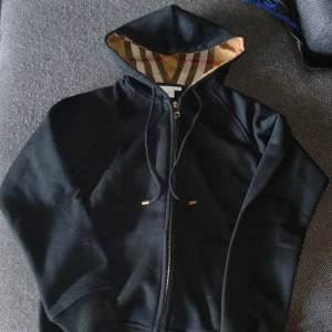 Svart Burberry hoodie med dragkedja - Svart hoodie från Burberry med klassiskt rutigt foder i huvan och guldfärgade detaljer på snörningen. Dragkedja framtill, broderad logga på bröstet och mjukt bomullsmaterial. Perfekt för dig som vill ha en stilren och lyxig streetwear-look.