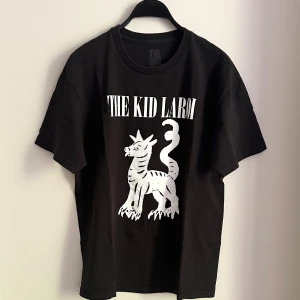 The Kid Laroi svart tour t-shirt - Svart t-shirt med vit tryck framtill av en tecknad drake och texten 'THE KID LAROI'. Baksidan har stor clown-illustration och turnéstäder från 2022. Klassisk passform och rund hals, perfekt för dig som gillar streetwear och musikmerch.Märkt storlek L Mått Längd ca. 70cm Bredd ca. 55cm