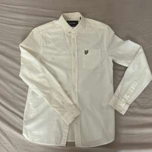 Vit skjorta från Lyle & Scott - Klassisk vit skjorta från Lyle & Scott med lång ärm och bröstficka med den ikoniska gula örnloggan. Skjortan har knappar hela vägen och en stilren, tidlös design i mjuk bomull. Perfekt för dig som gillar enkel och clean stil.