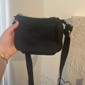  axelväska -  svart axelväska med justerbart band och dragkedja upptill. Väskan har en enkel, clean design och passar perfekt för att bära det viktigaste. Praktisk storlek och lätt att matcha med olika outfits.