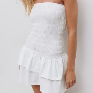 Vit axelbandslös kortklänning - Supersöt vit kortklänning med smockad överdel och volangkant nertill. Axelbandslös modell som ger en fräsch och somrig vibe. Perfekt för varma dagar när du vill känna dig både chill och snygg. Helt oanvänd endast testad passar dom som använder xs/s