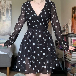 Svart omlottklänning med stjärnor från Glamorous - Supersöt svart kort omlottklänning från Glamorous med vita stjärnor över hela tyget. Klänningen har långa ärmar i tunt material, v-ringning och knytband i midjan. Perfekt för dig som gillar statement-plagg med coolt mönster och behöver en snygg klänning i sommar!