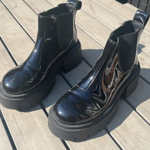 Svarta chunky chelsea boots lack - Säljer ett par svarta chelsea boots med chunky platåsula och glansig lackfinish. Skorna har elastiska paneler på sidorna och dragflik bak för enkel påtagning. Perfekta för dig som vill sticka ut med en edgy och trendig look.