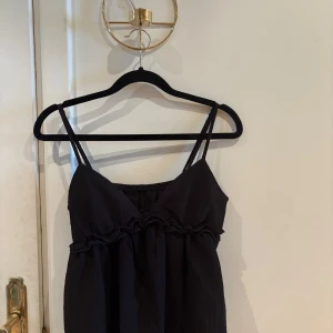 Svart linnetopp med volangdetalj - Säljer en svart linnetopp med tunna axelband och volangdetalj under bysten. Toppen har en lätt strukturerad yta och är luftig i modellen, perfekt för varma dagar. Enkel men ändå söt design som passar till mycket.