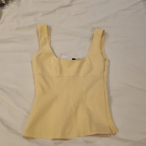 Gul korsett-topp från H&M Divided XXS - Supersöt ljusgul korsett-topp från H&M Divided i storlek XXS. Toppen har breda axelband, figurnära passform och dold dragkedja i sidan. Tillverkad i polyesterblandning med lite stretch för en snygg siluett. Perfekt för att lyfta vilken outfit som helst!