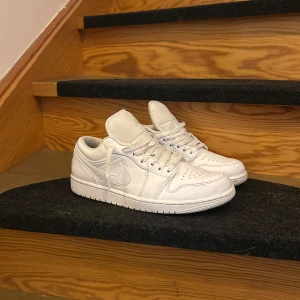 Nike Air Jordan 1 Low Triple White - Snygga Nike Air Jordan 1 Low i helvitt skinn med klassisk siluett och perforerad tå. Skorna har låga skaft, vit sula och diskreta broderade Air Jordan-loggor på hälen. Perfekta för dig som gillar clean och stilren look.  De kommer med box och crease skydd.