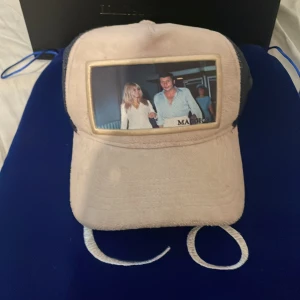 Maggiore keps med foto och mesh - Unik keps från Maggiore med beige front i plyschliknande material och svart mesh baktill. Framsidan har en inramad bild med motiv av två personer. Justerbar passform och cool kontrast mellan material och färger. Perfekt statement-piece för dig som vill sticka ut.