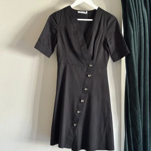 Svart omlottklänning  - Svart kortklänning från NA-KD med v-ringning och omlottkänsla. Klänningen har korta ärmar, dekorativa knappar framtill och lätt utställd kjol. Klänningen går att ha både med och utan medföljande bälte i samma tyg. Perfekt för dig som gillar stilrena och enkla plagg med snygga detaljer.