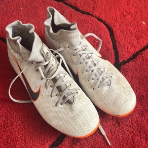 Vita Nike fotbollsskor med orange detaljer - Säljer ett par vita Nike fotbollsskor med högt skaft och orange detaljer. Skorna har snörning och en svart Nike-swoosh längs sidan. Storlek 40