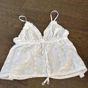 Vit broderad linnetopp från Censored - Supersöt vit linnetopp från Censored med smala axelband, v-ringning och volangkant. Toppen har broderade blommor, knyt i midjan och är lätt croppad. Perfekt för varma dagar