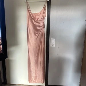 Ljusrosa långklänning med slits - Säljer en ljusrosa långklänning med tunna axelband och draperad urringning. Klänningen har en snygg slits på sidan och är gjord i ett glansigt, satinliknande material som ger en lyxig känsla. Perfekt för dig som vill sticka ut med en elegant och trendig look.
