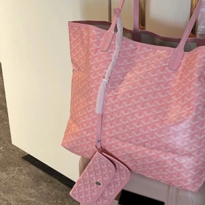 Rosa shoppingväska  - Rosa shoppingväska med ikoniskt mönster i vitt, samt  har två smala axelband och är rymlig nog för allt du behöver. Plånboken tillkommer och postar iväg samma dag.