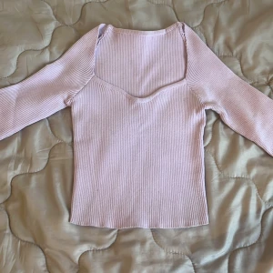 Ljusrosa ribbad långärmad topp - Supersöt ljuslila långärmad topp med ribbad struktur och snygg v-ringad halsringning. Sitter tajt och har en mjuk känsla, perfekt att styla med jeans eller kjol. Enkel men ändå trendig design som passar året runt.