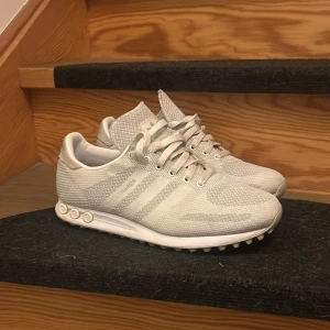 La Trainers - Snygga vita Adidas sneakers med diskret mönster och klassiska tre ränder på sidan. Skorna har snörning, platt sula och är tillverkade i ett lätt syntetmaterial som andas. Perfekta för dig som gillar stilrena och sportiga skor.
