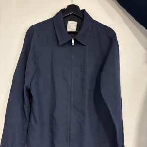 Blå jacka från Sandro paris - Snygg blå jacka från Sandro med dragkedja och en bröstficka. Perfekt för en stilren look. Jackan har långa ärmar och en klassisk krage. Jackan är i fint skick men några små defekter. Lite sliten på bröstfickan samt på ena ärmen, se bild 3&4. Hör av dig för fler bilder eller frågor!