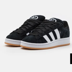 Svarta Adidas Campus sneakers - Snygga svarta Adidas Campus sneakers med klassiska vita ränder och gummisula. Skorna har en stilren design med snörning och är perfekta för en avslappnad stil. Den svarta färgen i kombination med de vita detaljerna ger en tidlös look.