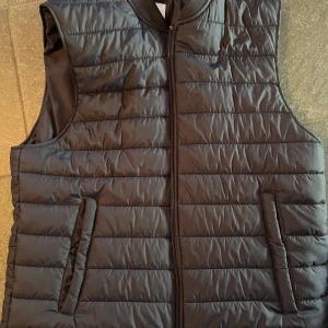 Svart quiltad väst från Zara - Snygg svart quiltad väst från Zara med dragkedja framtill. Perfekt för lager-på-lager under kyligare dagar. Västen har en stilren design med två fickor på sidorna.