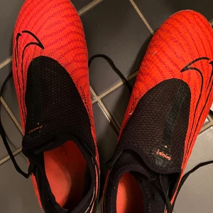  Röda Nike phantom GX  - Säljer ett par röda phantom Gx Mg ghost lace från Nike. Skorna har en strömlinjeformad design med dobbar för optimalt grepp på planen. Perfekta för fotbollsspelare som vill ha ett par bekväma och lätta skor. Skosnörena är under flärpen perfekt för dig som hatar att ständigt behöva knyta om dina skor på planen. Skorna är i bra skick. Skriv för frågor.