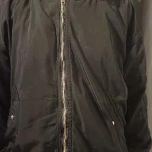 Svart bomberjacka med dragkedja - Snygg svart bomberjacka med dragkedja framtill och ribbade muddar. Jackan har en klassisk design med två fickor med knappar. Perfekt för en cool och avslappnad stil.