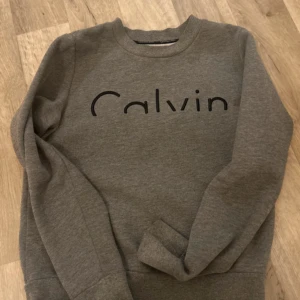 Grå sweatshirt från Calvin Klein - Säljer en stilren grå sweatshirt från Calvin Klein med texten 'Calvin' på bröstet. Tröjan har långa ärmar och en bekväm passform. Perfekt för en avslappnad look.