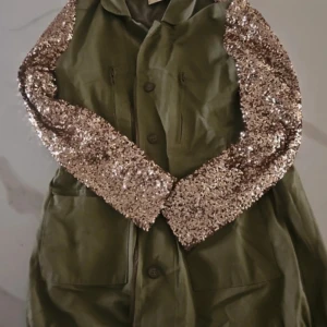 Olivgrön jacka med paljettärmar från legende angels - Snygg olivgrön jacka med glittriga paljettärmar som verkligen sticker ut. Jackan har knappar framtill och stora fickor, perfekt för att ge en cool touch till din outfit. Den är långärmad och har en unik design som kombinerar militärstil med glamour.