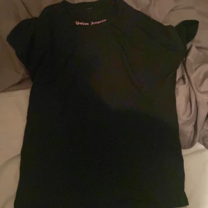Svart t-shirt från Palm Angels - Säljer en svart t-shirt från Palm Angels med deras ikoniska logga i vitt och rosa på ryggen. T-shirten har korta ärmar och en normal passform. Perfekt för en stilren look.