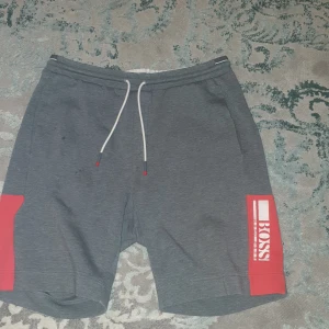 Grå shorts från Hugo Boss äkta tror jag - Snygga grå shorts från Boss med röda detaljer och logga på sidan. De har en bekväm passform med snörning i midjan och är perfekta för en avslappnad stil. Materialet känns mjukt och skönt, vilket gör dem idealiska för både vardag och fritid.