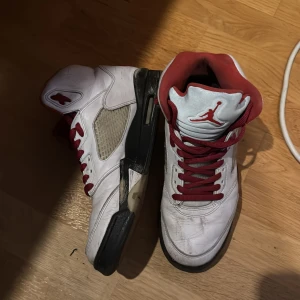 Jordan 5 Retro Fire Red Silver Tongue - Säljer ett par Jordan 5 Retro Fire Red Silver Tongue från 2020. Skorna är vita med röda och svarta detaljer samt har en ikonisk design med snörning och hög ankel. Perfekta för sneakerheads som vill ha en klassisk stil.