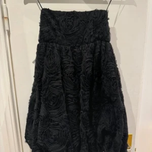 Svart klänning med rosdetaljer från Festive Days - Snygg svart klänning  från Festive Days med unika rosdetaljer som ger en lyxig känsla. Perfekt för speciella tillfällen. Kjolen har en voluminös design och är i storlek S. Ett måste för garderoben!