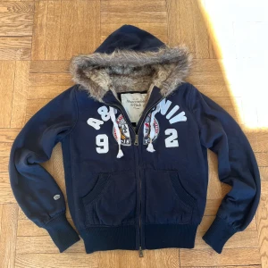 Mörkblå hoodie från Abercrombie & Fitch - Säljer en mörkblå hoodie från Abercrombie & Fitch med pälskantad huva och dragkedja. Den har vita tryck och broderier på framsidan samt ribbade muddar. Storlek m men passar som s