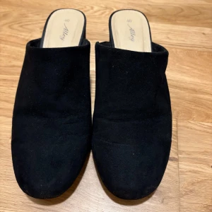 Svarta mocka slip-ins från Alley - Snygga svarta slip-ins i mocka från Alley. Dessa skor har en stilren design med en låg klack och en bekväm passform. Perfekta för en avslappnad men elegant look.