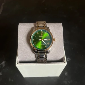 Grön klocka med silverarmband - Snygg klocka med en iögonfallande grön urtavla och silverfärgat armband. Klockan har en kvartsrörelse och visar både dag och datum. Perfekt för den som vill ha en stilren accessoar med en färgklick. Tar emot byte!!