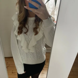 Vit stickad tröja med volangdetaljer - Säljer en stilren vit stickad tröja med vackra volangdetaljer framtill. Tröjan har ett klassiskt flätmönster och långa ärmar, perfekt för kyligare dagar. Passar både till jeans och kjol för en chic look.