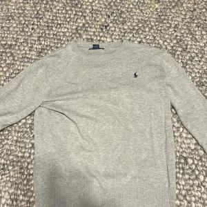 Grå tröja från Polo Ralph Lauren - Säljer en stilren grå tröja från Polo Ralph Lauren med deras ikoniska logga broderad på bröstet. Tröjan har långa ärmar och ribbade muddar vid ärmslut och nederkant. Perfekt för en klassisk och avslappnad look.