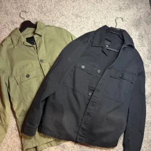 Svart och grön overshirt  - Två stilrena overshirts från 157 i svart och grön. Båda har långa ärmar och knappar framtill samt två bröstfickor med knappstängning. Perfekta för lager-på-lager-stil under höst och vår. Skick 10/10, storlek S. Kan sälja styckvis eller 2-pack beroende på överenskommelser. Pris kan diskuteras vid snabb affär! 
