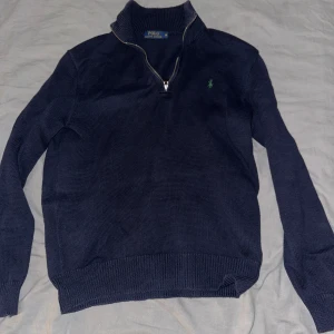 Mörkblå tröja från Polo Ralph Lauren - Säljer en stilren mörkblå tröja från Polo Ralph Lauren med dragkedja vid halsen och ett broderat grönt märke på bröstet. Tröjan har långa ärmar och ribbade muddar vid ärmslut och nederkant. Perfekt för en klassisk look.