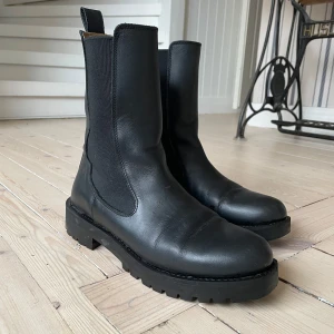 Svarta boots i skinn - Snygga svarta boots i skinn med en robust sula och elastiska paneler på sidorna för enkel på- och avtagning. Perfekta för en stilren look. Lite högre i skaftet och de är inköpta på Other Stories. Storlek 40.