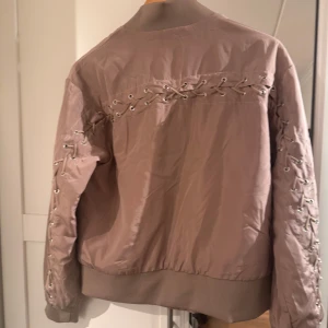 Beige bomberjacka med snörning - Snygg beige bomberjacka från NA-KD med unik snörningsdetalj på ryggen och ärmarna. Jackan har en dragkedja framtill och ribbade muddar vid ärmslut och nederkant. Perfekt för en trendig look!