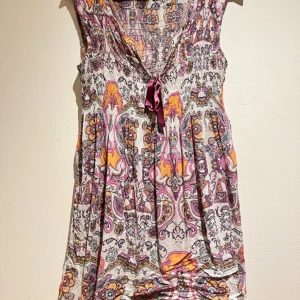 Mönstrad klänning från Odd Molly - Säljer en färgglad klänning från Odd Molly med ett livfullt mönster i rosa, orange och lila toner. Klänningen är ärmlös och har en v-ringad design med knytning framtill. Perfekt för en bohemisk look!